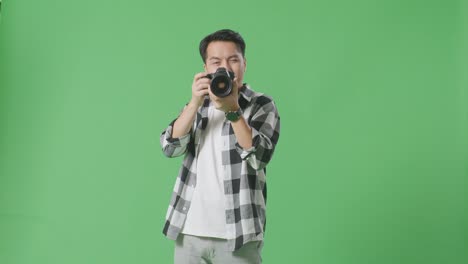 asian photographer using a camera taking pictures and smiling touching his chest while standing on green screen background in the studio