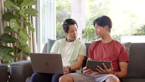 Asian father and teenage son sit on couch using a laptop and tablet respectively