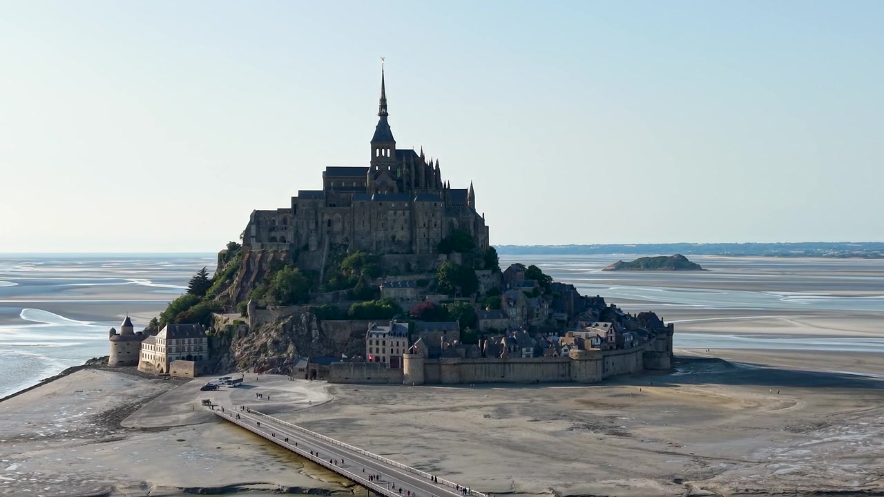 Vista aérea aproximada de drone do Mont Saint-Michel, destacando a icónica abadia medieval, a vila fortificada, as antigas muralhas e as planícies de maré circundantes na maré baixa