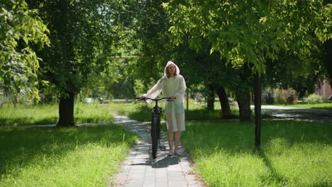 young woman in translucent raincoat strolls along a sunny pathway with bicycle, stopping to place stand, remove hood, and lean on handlebars, background features lush greenery and trees