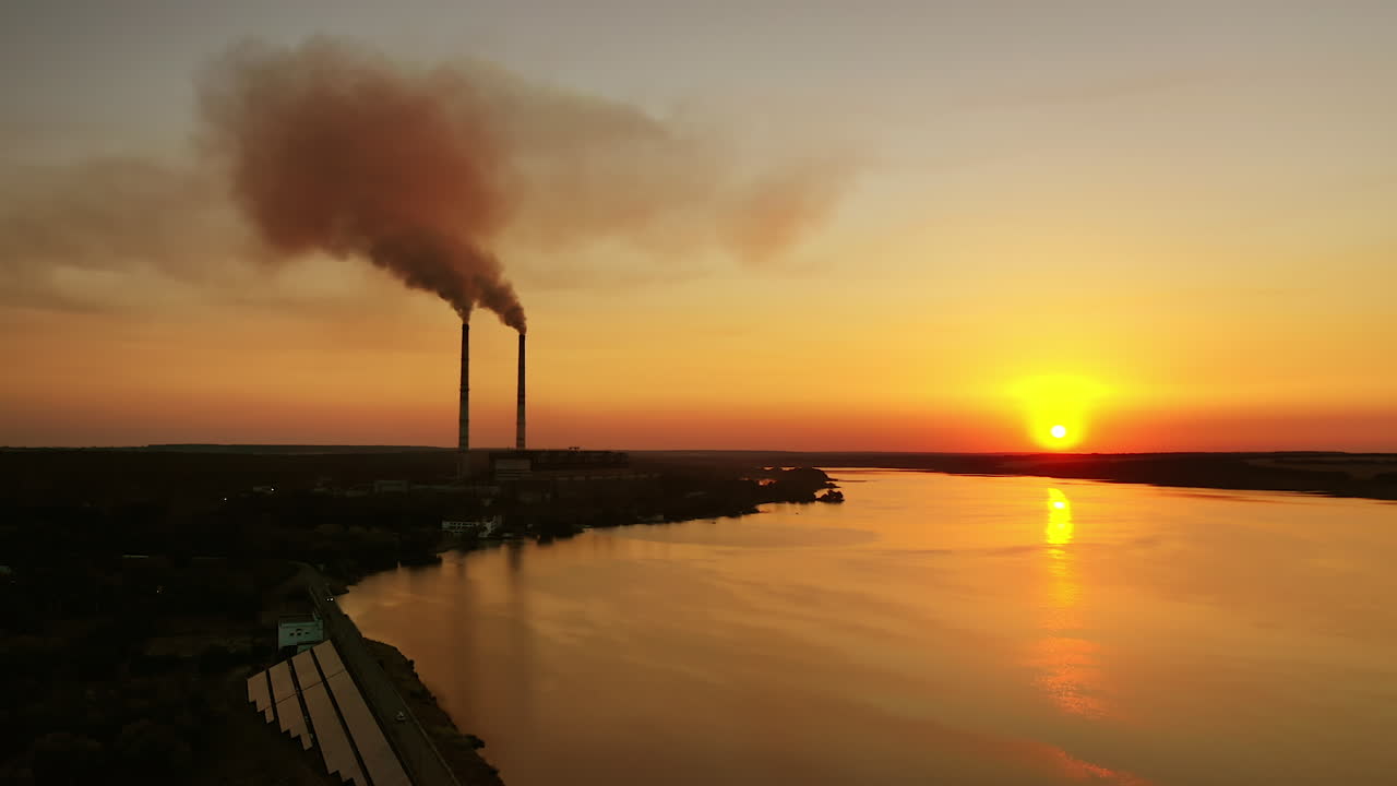 Orange sunset over the water. Harmful plant with industrial pipes near the river. Dirty emissions pollute the air in the evening.