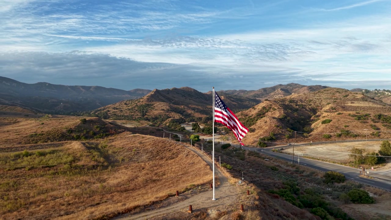 Sapuan udara yang indah di sekitar Bendera Amerika yang berkibar anggun. Saat drone meluncur perlahan dari satu sisi ke sisi lain, pemandangan terbuka ke pegunungan dan langit biru yang mengelilingi Bendera Amerika.