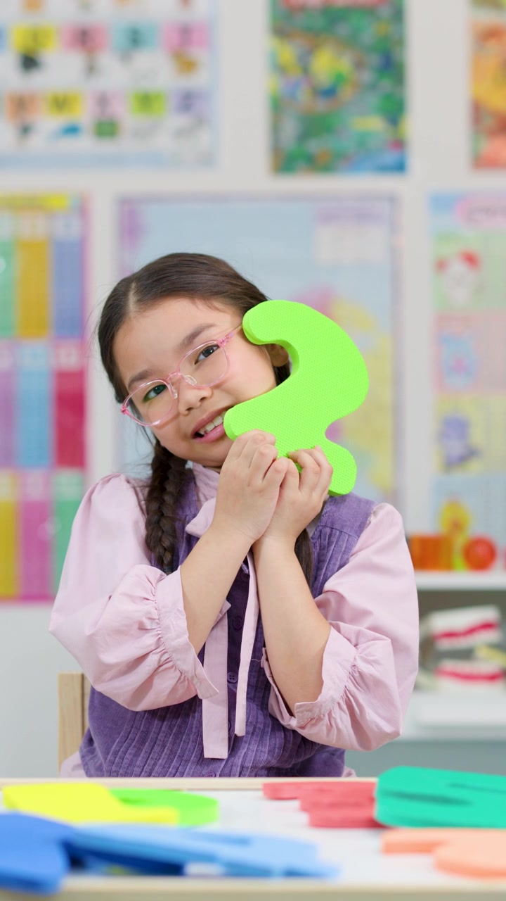 Una ragazza asiatica con gli occhiali seleziona e mostra il numero due in gommapiuma in un'aula colorata e ben illuminata.