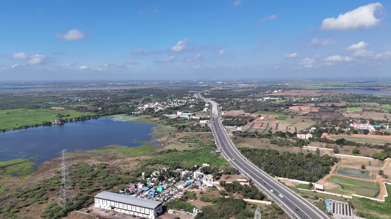 A modern highway with busy traffic spans Chennai's urban-rural landscape from above. Residential areas, green fields, and distant towns are visible under a clear, bright sky