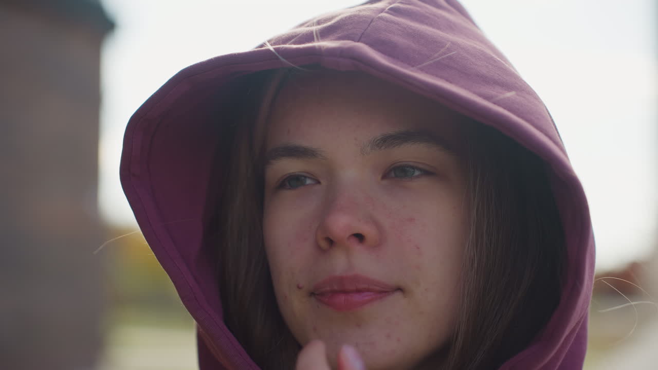 Hoodie wearing athlete hands in pocket walking elegantly adjusting hoodie while breeze blows hair under sunny sky on gray tiled pavement with blurred urban park backdrop capturing fluid style motion