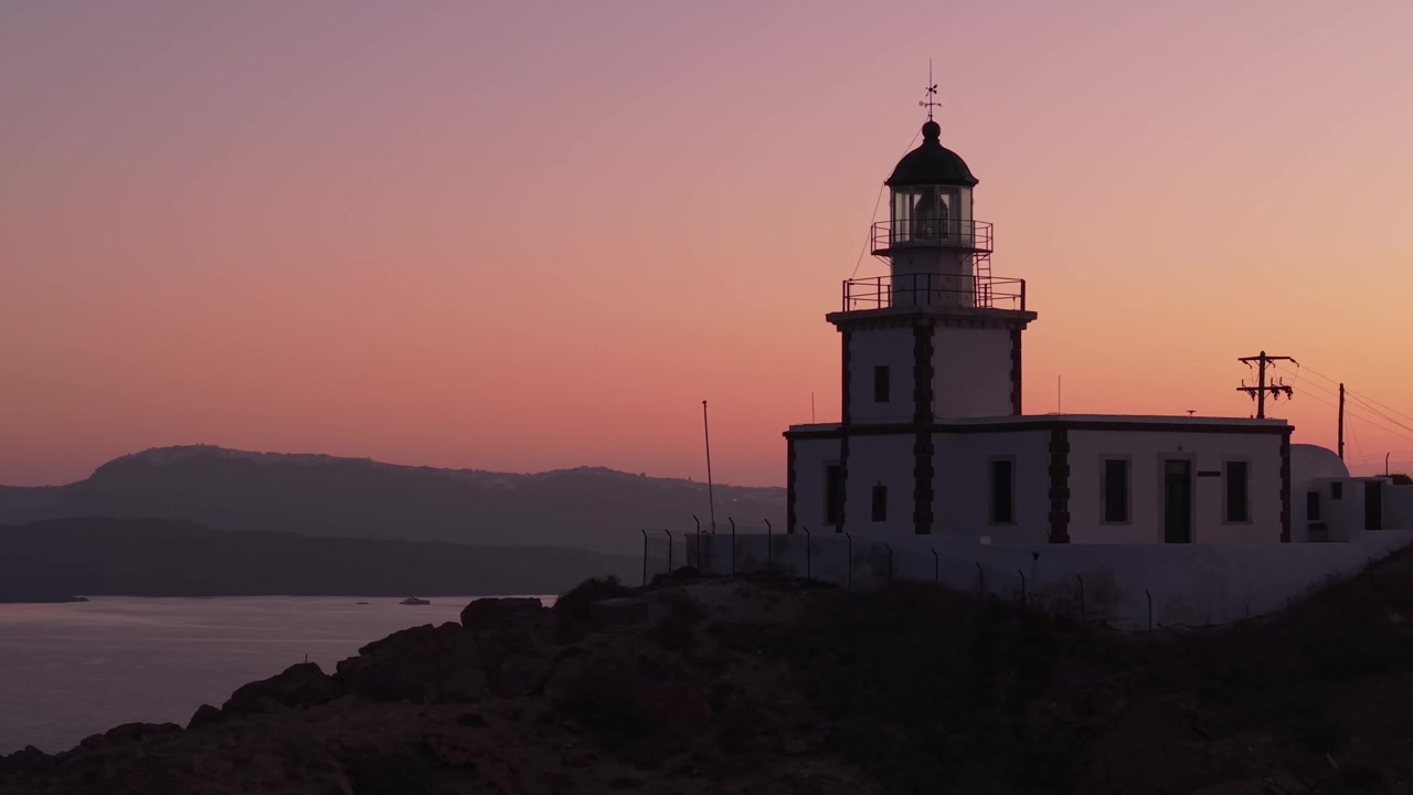 Drone in orbita attorno al faro di Santorini all'alba, che cattura un'atmosfera serena con luce tranquilla, le scogliere della caldera e il pacifico Mar Egeo.