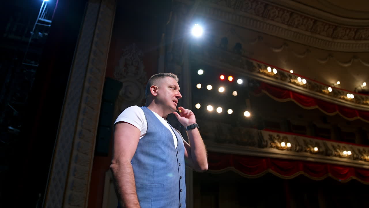 Smiling actor on stage. Middle-aged man standing on the scene of a theater and talking to the audience. Acting at bright spotlights. Side view.