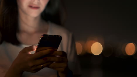 at night a young girl sits on the grass in a big city and looks into the smartphone screen writes a message and maintains her personal blog.