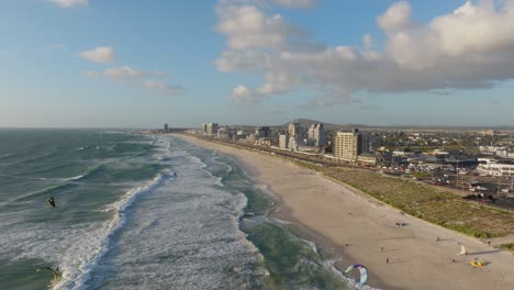 drone captures bloubergstrand’s wide sandy beach and powerful atlantic ocean surf lined with kite surfers and modern buildings, looking toward cape town on a breezy clear day