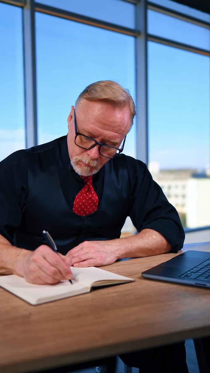 Serious businessman in black clothes and red tie sits at desk in the office. Man having some troubles, fixes his tie nervously and takes notes. Vertical video.