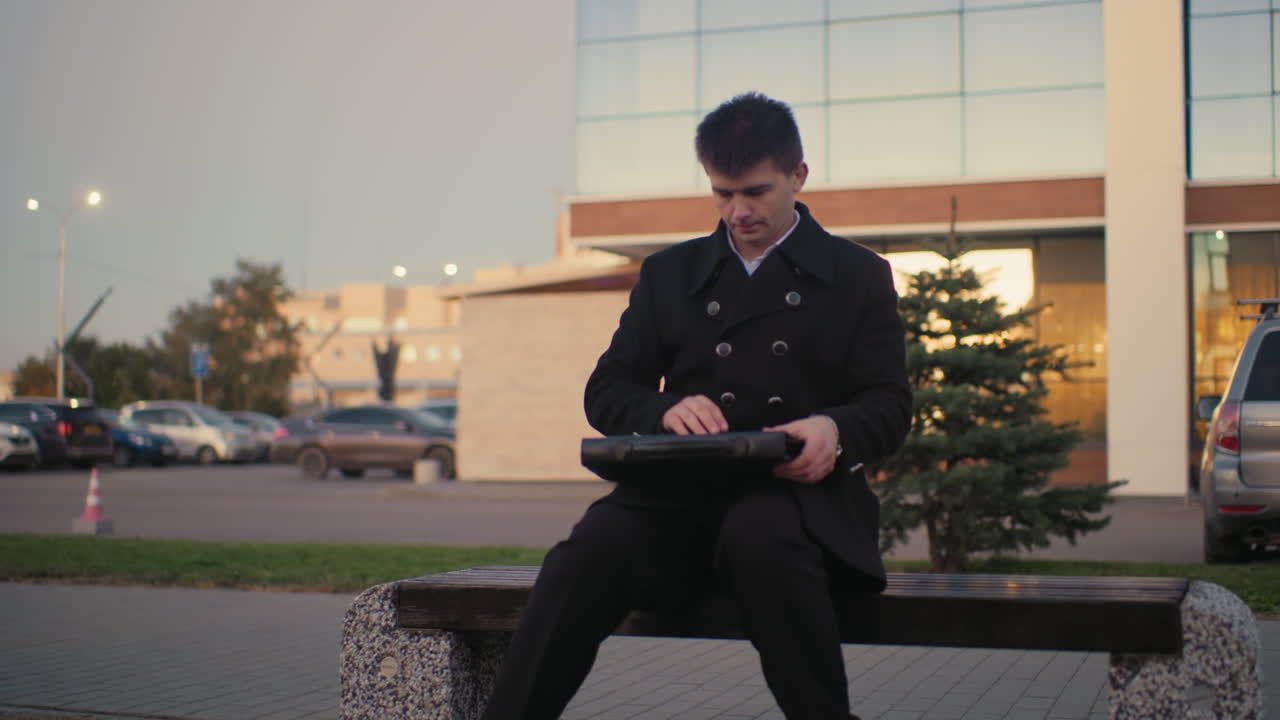 검은 옷을 입은 남자가 벤치에 다가와, 현대적인 건물과 주차된 차와 함께 야외 환경에서 브리프케이스를 열고, 새벽에 배경에서 움직이는 차, 이른 아침에 빛나는 거리 조명