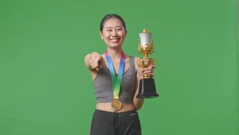 asian woman with a gold medal and trophy touching her chest then smiling and pointing to camera being proud of herself on green screen background in the studio