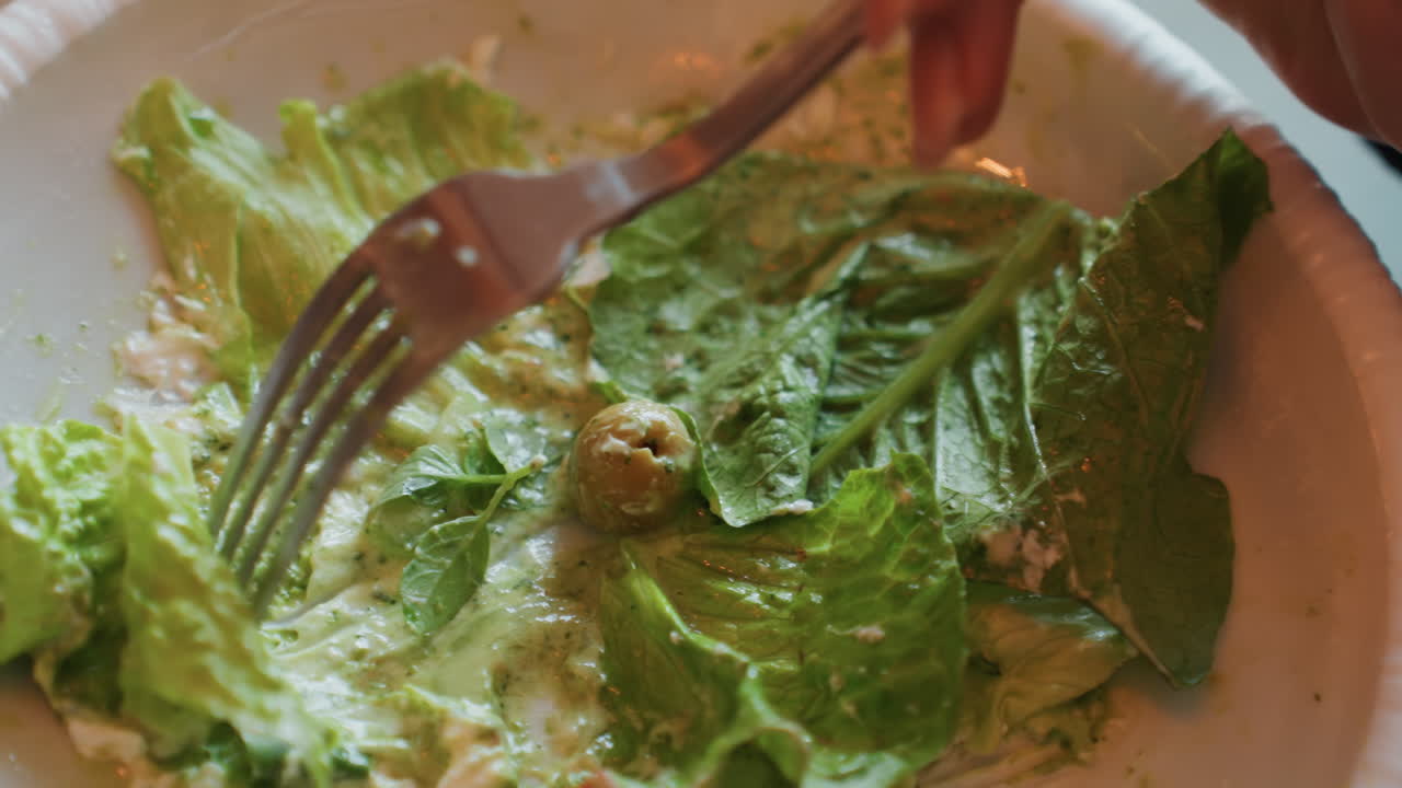 Primo piano di insalata avanzata su un piatto di carta con una forchetta che raschia il condimento cremoso, una singola oliva appoggiata vicino a foglie di lattuga romana, luce calda della cucina, piatto usa e getta implica un pranzo veloce a casa, rilassato e solitario