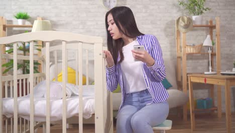 young asian woman with a smartphone cradles a baby in a crib sitting in the living room