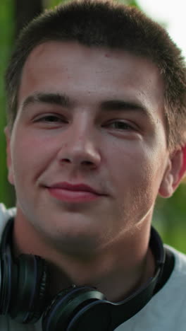 close-up of man with black headphones around neck, wearing white top and seated outdoors with warm smile, surrounded by green trees and soft outdoor lighting