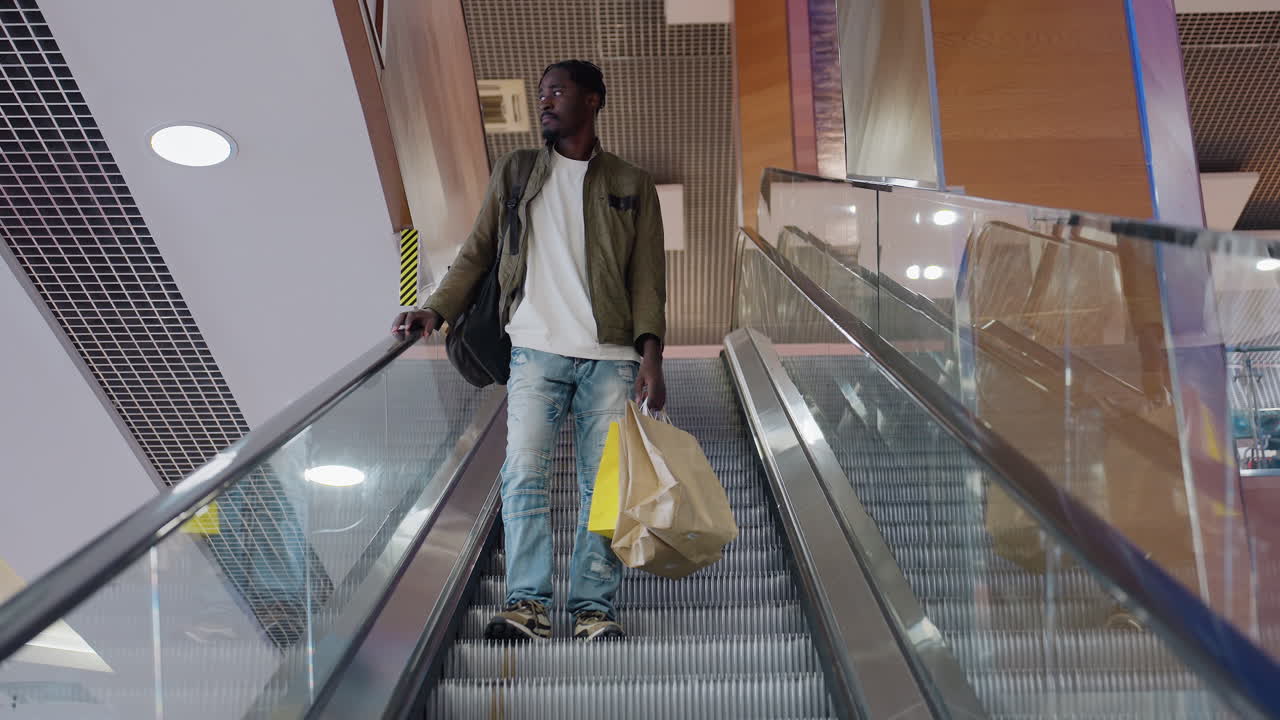 Young student holding paper shopping bags descends escalator inside modern shopping mall while looking around in relaxed posture wearing casual outfit in spacious bright retail environment