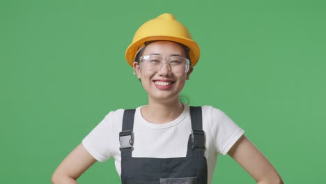 close up of asian woman worker wearing goggles and safety helmet standing with arms akimbo looking at camera and smiling in the green screen background studio