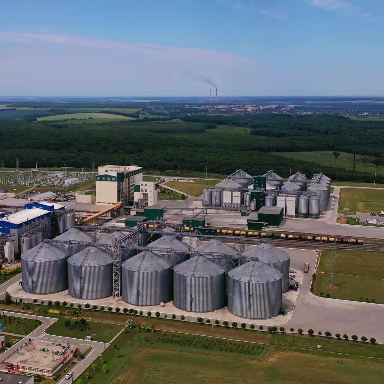 Silver granaries at huge modern plant. Abundant industrial territory with railways in the middle. Aerial perspective. Farmlands backdrop