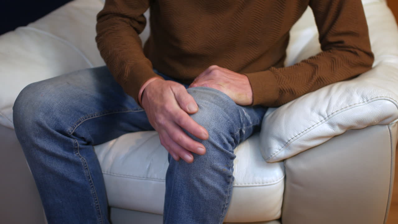 A man sitting on a white couch clutching his knee with both hands indicating localized pain or discomfort Close up view highlighting the gesture against a casual home setting