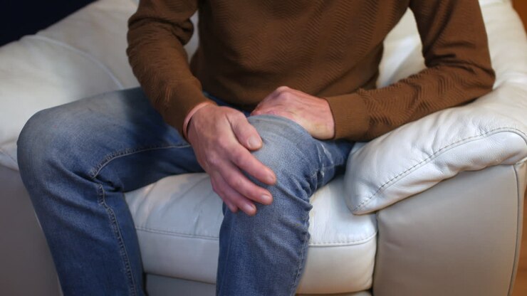 A man sitting on a white couch clutching his knee with both hands indicating localized pain or discomfort Close up view highlighting the gesture against a casual home setting