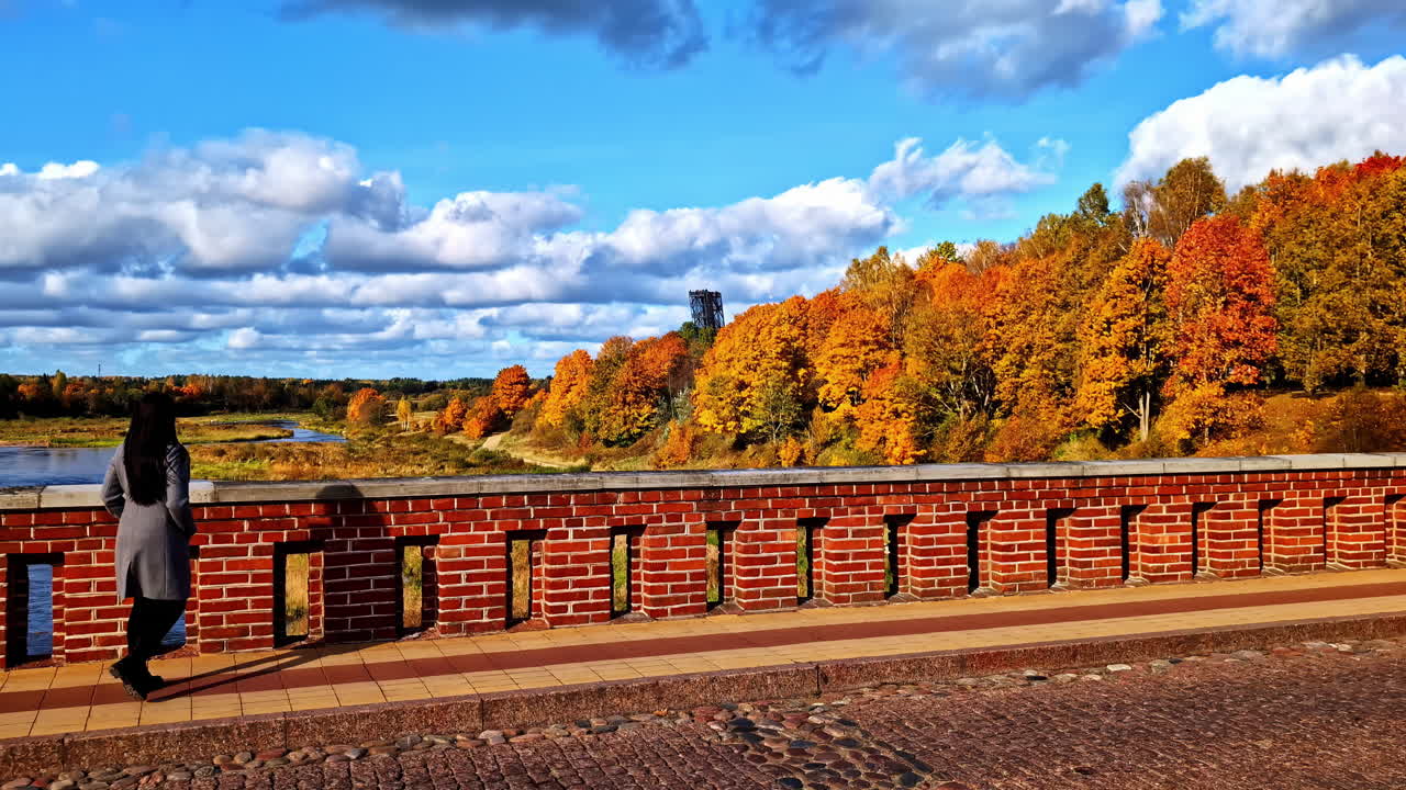 A dark-haired woman stands on a bridge in autumn sunshine, surrounded by vibrant nature and colorful leaves.