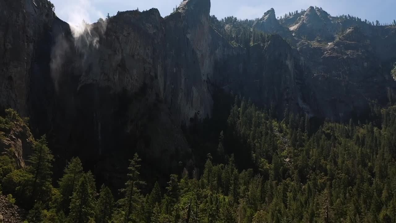 Aerial view of a tall waterfall streaming down steep granite cliffs surrounded by dense pine forest and rugged mountain peaks in Yosemite National Park, California