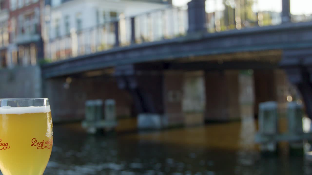 Hand lifts beer glass beside canal in Haarlem, Netherlands, with blurred bridge in daylight