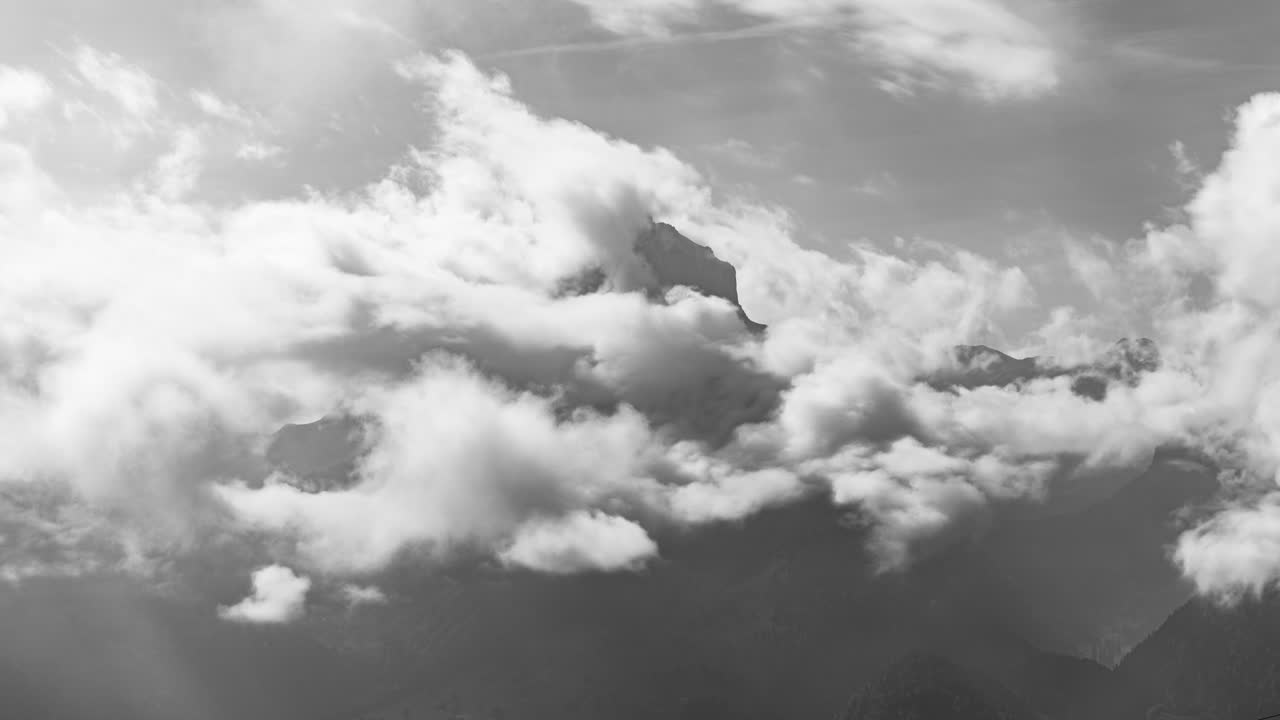A black-and-white timelapse of dramatic clouds rolling over the Swiss Alps in Amden, St. Gallen, Switzerland. Shadows and light interact, creating a powerful visual.