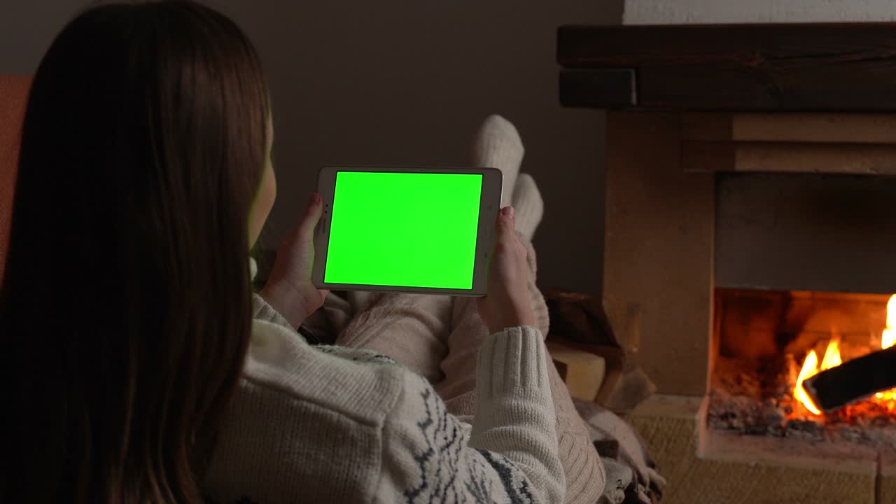 mujer acostada en el sofá frente a la chimenea sosteniendo una pantalla verde de tableta, viendo la pantalla clave de croma junto a la chiminea