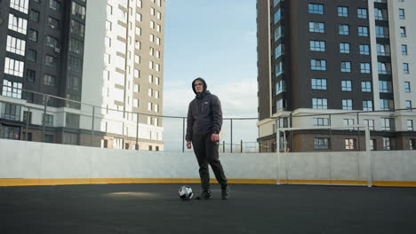 sportsman in hoodie and athletic wear practicing ball control, standing on sport arena field with goalpost behind, urban background highlights tall modern office buildings