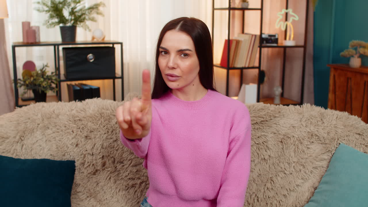 Young woman sitting on sofa waving finger in front expressing disagreement rejection no disapproval
