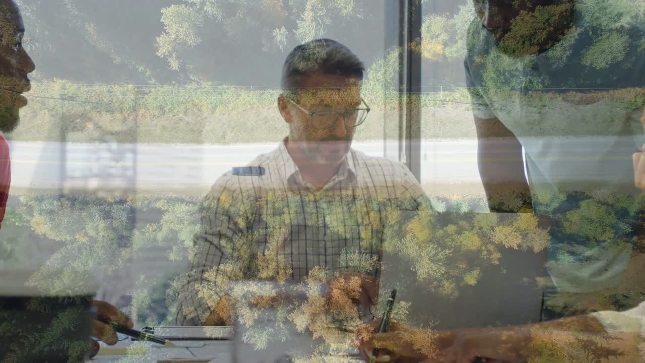 Responsabile aziendale che prende appunti mentre un collega indica un laptop, iniziando la revisione di un documento sotto una sovrapposizione di fogliame.