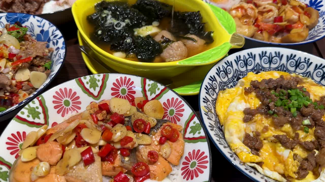 A hand uses a spoon to serve seaweed soup from a yellow bowl, surrounded by colorful Thai dishes under bright indoor lighting, with close-up camera movement