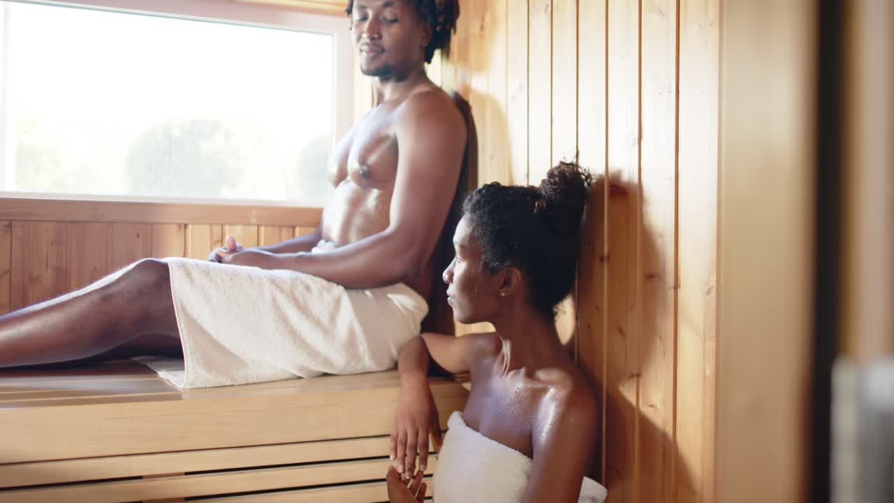 Casal afro-americano reagindo à câmera se afastando, compartilhando um momento particular na sauna usando toalhas.