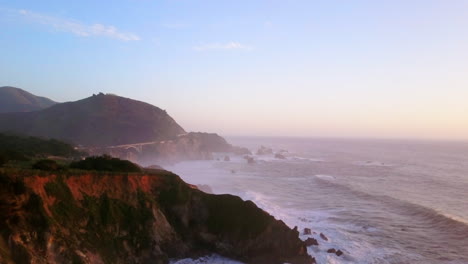 bixby creek bridge big sur california vuelo aéreo cinematográfico de drones océano pacífico ni cal invierno verano gran ola de oleaje chocando costa accidentada tarde hora de oro puesta de sol rosa movimiento hacia atrás