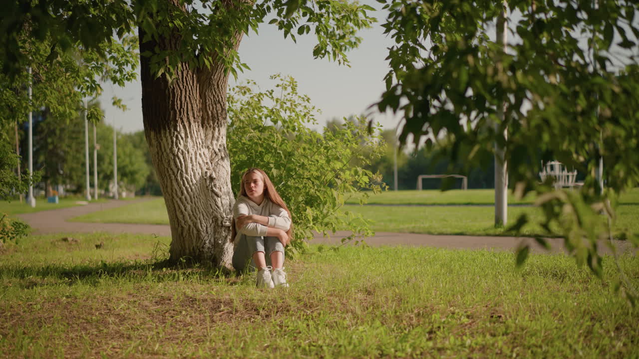 mujer se sienta en el suelo cubierto de hierba bajo un árbol con expresión pensativa, cruzando los brazos sobre las piernas, la luz del sol se refleja en su cara mientras mira hacia la derecha, el fondo incluye postes y estadio