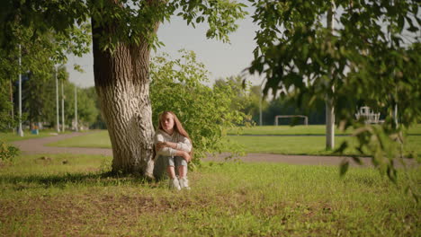 woman sits on grassy ground under tree with thoughtful expression, folding arms over legs, sunlight reflecting on her face as she looks to the right, background includes poles and stadium