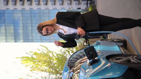 A professional woman in a business suit charges her electric car. She stands next to a blue electric car, highlighting ecofriendly transportation and modern technology