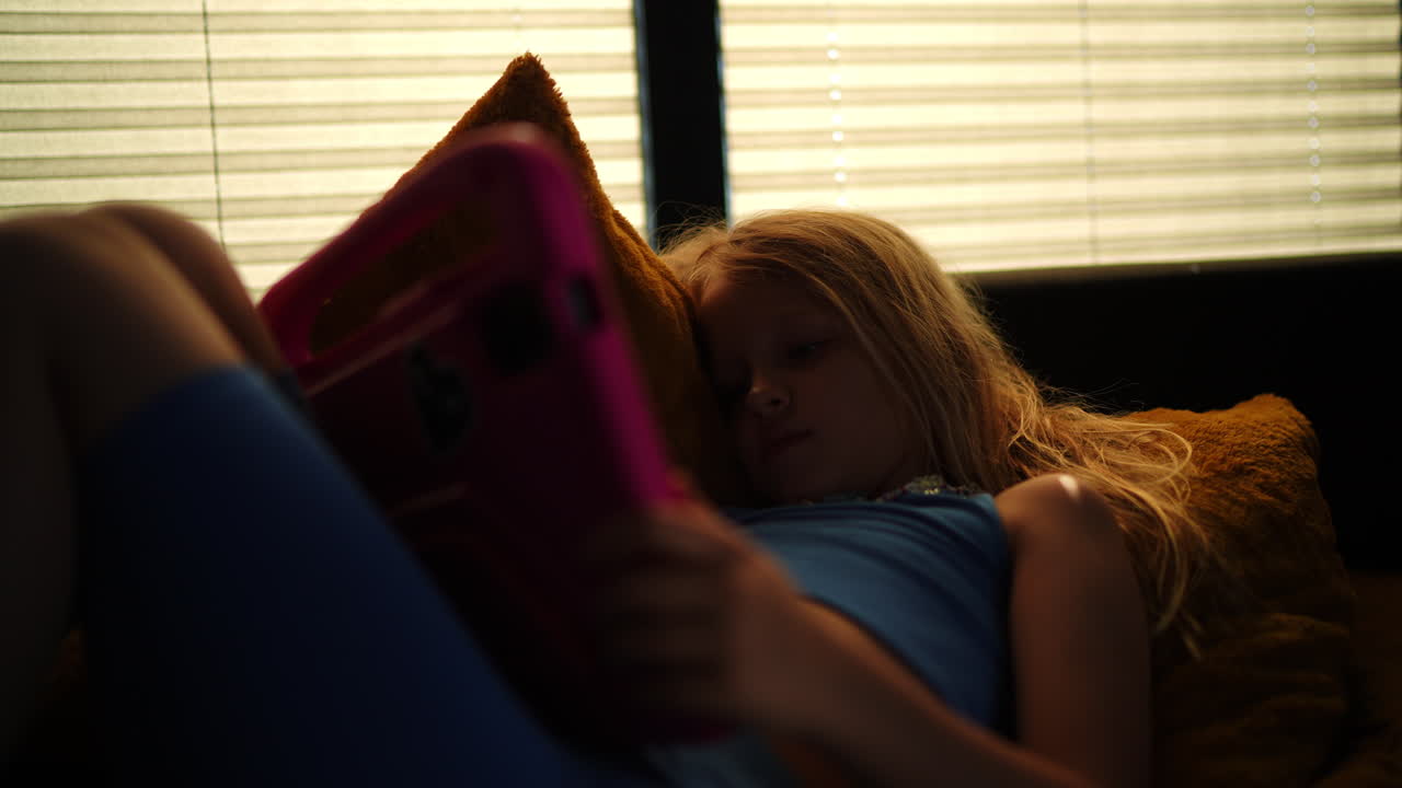 Blond haired girl relaxing comfortably on a a sofa, playing tablet near sunlit window with Venetian blinds, enjoying quiet leisure moment