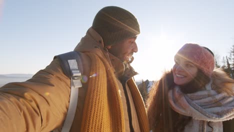 Feliz-Pareja-Intercultural-Joven-En-Ropa-De-Invierno-Saludando-A-La-Cámara-De-Pie-Contra-El-Cielo-Azul-Sobre-Montañas-Cubiertas-De-Nieve-Durante-El-Frío-En-Invierno