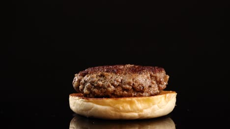 a single beef burger patty on a plain bun rotates smoothly on a glossy black surface, illuminated by dramatic studio lighting with a dark backdrop