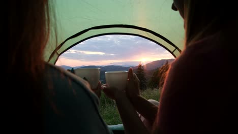 two people were lying in a tent overlooking the mountains young girls drinking hot tea on top of a hill overlooking the sunset
