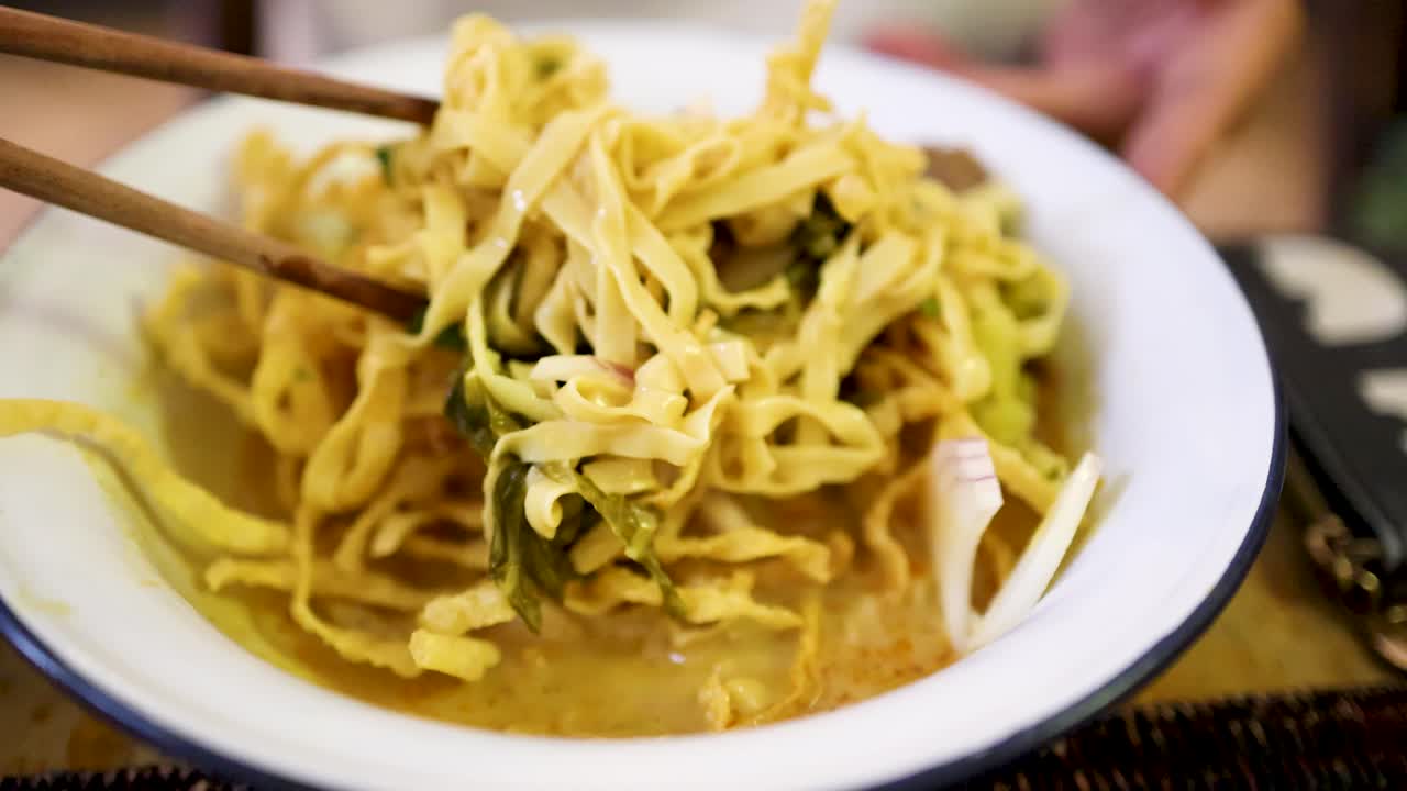 Hand uses chopsticks to lift yellow curry noodles with beef and vegetables from a white bowl in bright, natural light at a Bangkok eatery