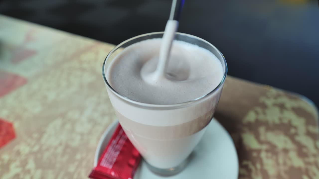 A spoon gently stirs frothy hot cocoa in a glass, creating a smooth swirl, Cafe