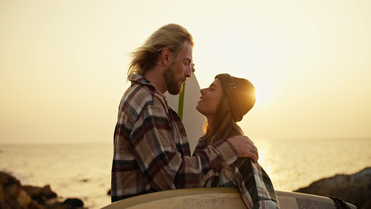 Happy blond guy with a beard in a checkered shirt kisses his blonde girlfriend, who holds a surfboard and rejoices on a rocky shore near the sea. Romantic meeting of surfers, guy and girl on a date in the morning