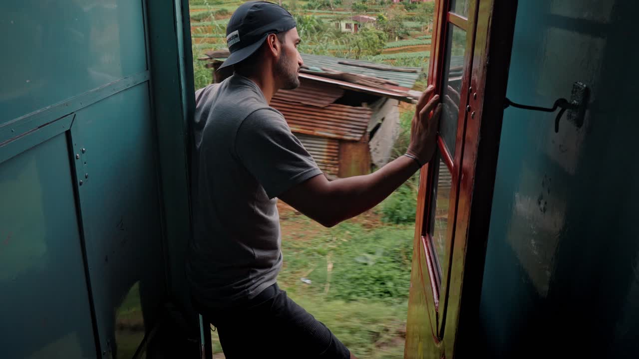 En mand sidder i den åbne dør på et kørende tog langs den naturskønne jernbanestrækning fra Ella til Kandy i Sri Lanka og kigger ud over de frodige grønne bakker, teplantager og den tropiske jungle.