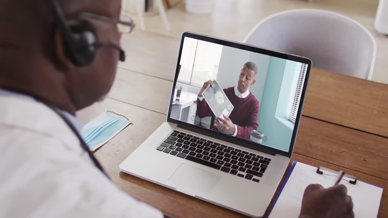 Ein afroamerikanischer Arzt mit Telefon-Headset macht sich Notizen während eines Videoanrufs am Laptop.