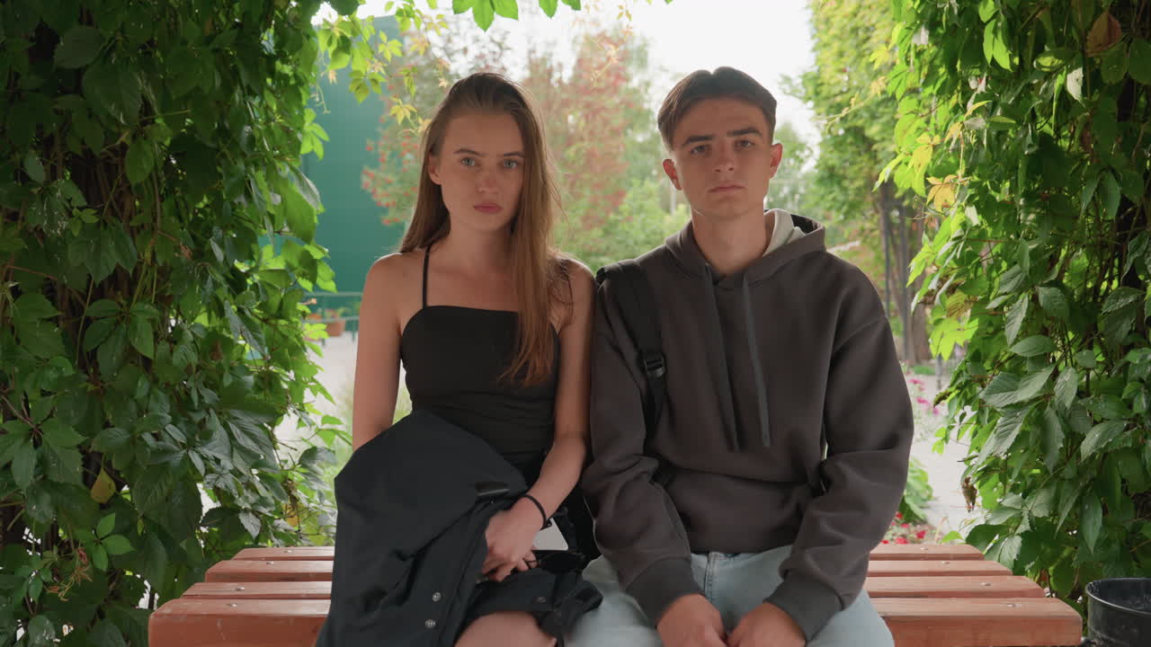 Due adolescenti seduti su una panchina sotto un arco frondoso; la ragazza con un abito nero e il ragazzo con una felpa grigia condividono uno sguardo silenzioso e intenso. La retroilluminazione naturale filtra attraverso la fitta vegetazione, primi piani spontanei catturano un’aria pensierosa.
