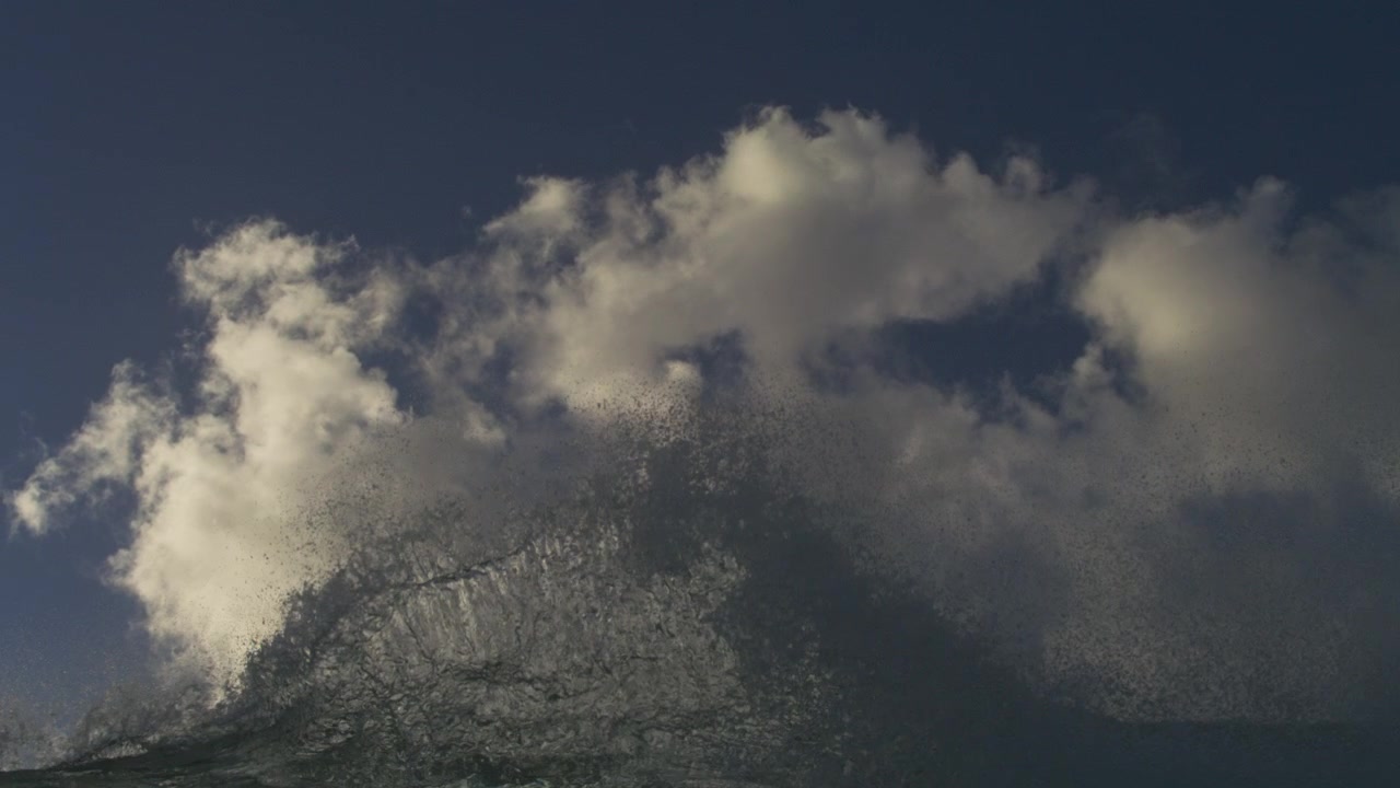 White spray erupting from an ocean wave beneath towering clouds in ultra slow motion