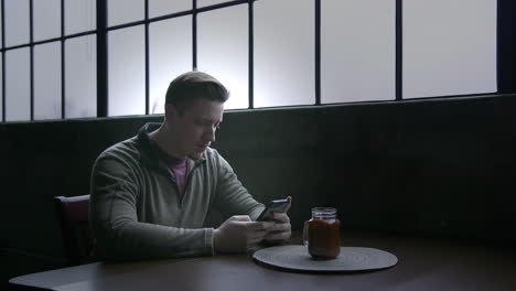 a young man sits at a table in his loft texting on a smartphone